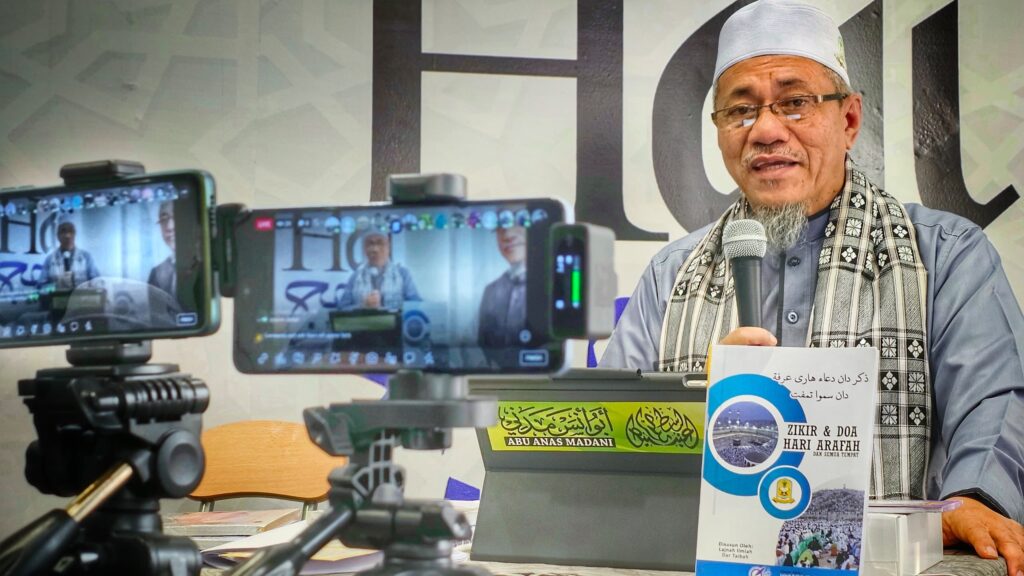 Dr. Abu Anas Madani menyampaikan kuliah sambil memegang mikrofon, dirakam oleh dua telefon pintar di atas tripod, dengan risalah Zikir & Doa Hari Arafah di atas meja